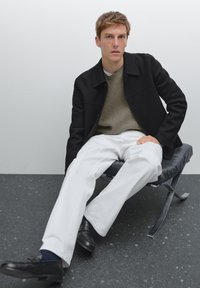 Sort frakke, olivengrøn ribbet sweater, hvide bukser, sorte loafers. Model sidder på en sort lænestol mod en neutral baggrund.
