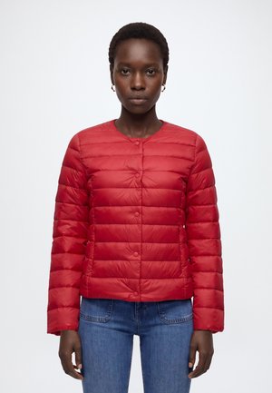 Femme portant une veste matelassée rouge à boutons et un jean bleu, debout devant un fond blanc uni.