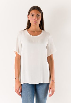 Donna con lunghi capelli scuri che indossa una maglietta bianca semplice, larga, a maniche corte e jeans blu, in piedi davanti a uno sfondo bianco.