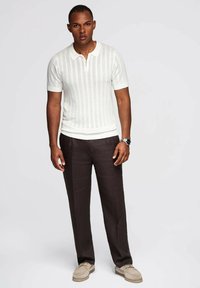 Camisa de polo de punto blanca, de manga corta, con rayas verticales, combinada con pantalones marrones oscuros. Zapatos color beige completan el conjunto.