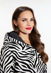 Pufová bunda se zebřím potiskem v černobílé barvě s kapucí, se lesklým povrchem a prošívaným designem. Modelka má dlouhé, vlnité vlasy.
