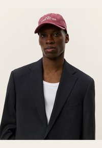 Bordeaux värvi müts, millel on tekst "LOWER WEST SIDE", kantud tumeda bleiseri all valge soonikkoes topi peal. Sile materjal, struktureeritud disain, mugav, kuid samas viimistletud.