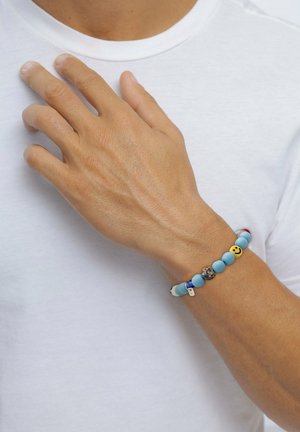 Bracelet en perles bleues comportant des perles de différentes couleurs, y compris un visage souriant doré et des accents à motifs, sur une main portant une chemise blanche.