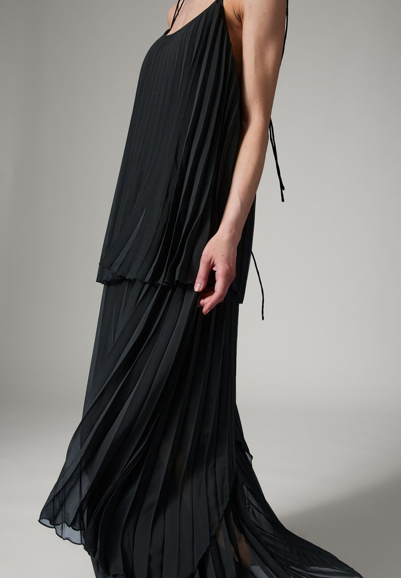 Femme portant une robe noire plissée sans manches avec des fines bretelles, debout devant un fond gris uni.
