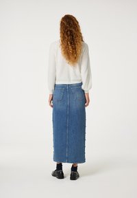 Denim maxi rok in het blauw met een rechte vorm, twee achterzakken en zijknopen. Gecombineerd met een witte gebreide trui en zwarte schoenen.