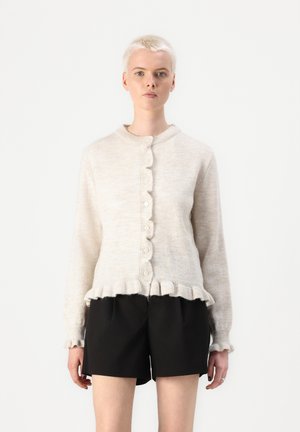 Lys beige cardigan med bølget kant og mansjetter, med strukturert strikk, rund krage og knapplukking foran. Kombinert med svarte shorts.