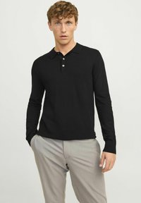 Camisa polo negra de manga larga en tejido suave, que presenta un cuello con solapa y una botonera de tres botones, combinada con pantalones grises claros.