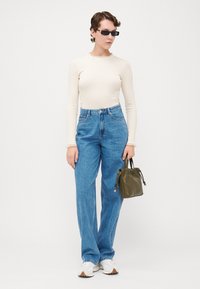 Haut crème à manches longues avec un col à volants, jeans bleu taille haute, baskets blanches et un petit sac vert olive. Style minimaliste.