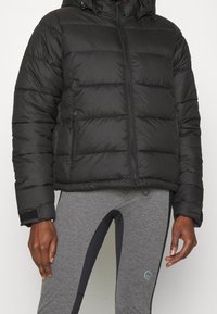 Chaqueta de plumas negra con capucha, con un diseño acolchado, cierre frontal y dos bolsillos laterales. Llevada sobre leggings grises con acentos oscuros.