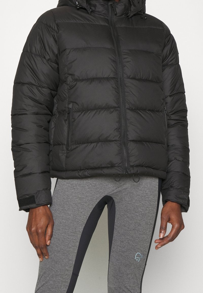Chaqueta de plumas negra con capucha, con un diseño acolchado, cierre frontal y dos bolsillos laterales. Llevada sobre leggings grises con acentos oscuros.