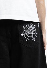 Black denim shorts featuring a detailed white spider and web embroidered design on the back pocket; tag with "WASHED" visible.