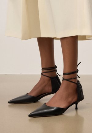 Tamnopute noge u crnim kitten cipelama sa špicastim vrhom i trakama koje se vežu oko gležnja, uparene s kremasti suknjom srednje dužine.