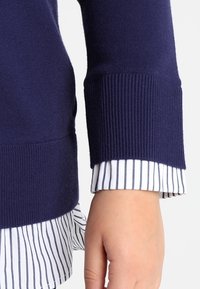 Marineblauer Pullover mit gerippten Bündchen, über einem weißen Hemd mit vertikalen blauen Streifen getragen. Nahaufnahme der Ärmeldetails und Stofftextur.