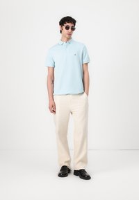 Camisa polo de color azul claro con mangas cortas y logo, combinada con pantalones de pierna ancha crema y zapatos negros. Modelo lleva gafas de sol.
