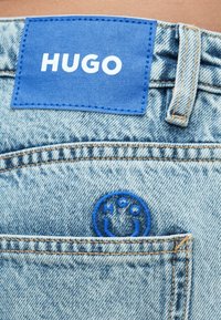Világoskék farmernadrág kék bőr címkével, amelyen a "HUGO" felirat szerepel, valamint kék hímzett logó a hátsó zseben. Látható varrási részletek.