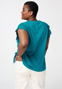 Blouse en teal avec des finitions en dentelle sur les manches, un col rond et une coupe décontractée. Associée à un pantalon clair, doté de poches et de détails au dos.
