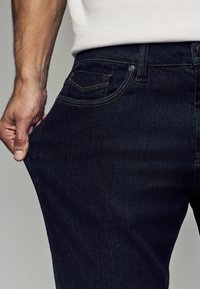 Jeans de denim oscuro con un diseño ajustado. Presenta un estilo clásico de cinco bolsillos y costuras en contrate a lo largo de las costuras.