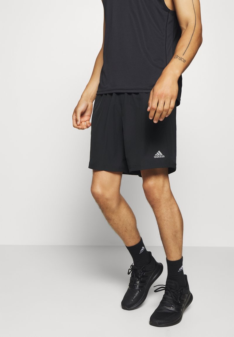 Personne portant un short de sport Adidas noir, un débardeur noir assorti, des chaussettes noires et des baskets noires, debout sur un fond clair uni.