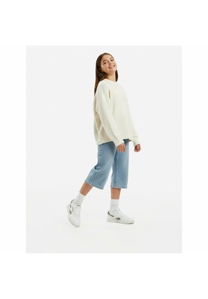 Mladá žena se usmívá, má na sobě nadměrný krémový svetr, krátké světle modré džíny, bílé ponožky a bílé tenisky, stojí na jedné noze na bílém pozadí.