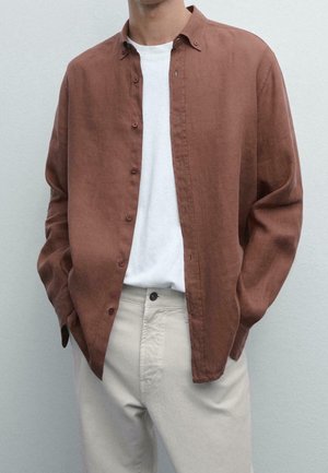 Personne portant une chemise marron à manches longues déboutonnée sur un t-shirt blanc avec un pantalon beige clair, les mains dans les poches.