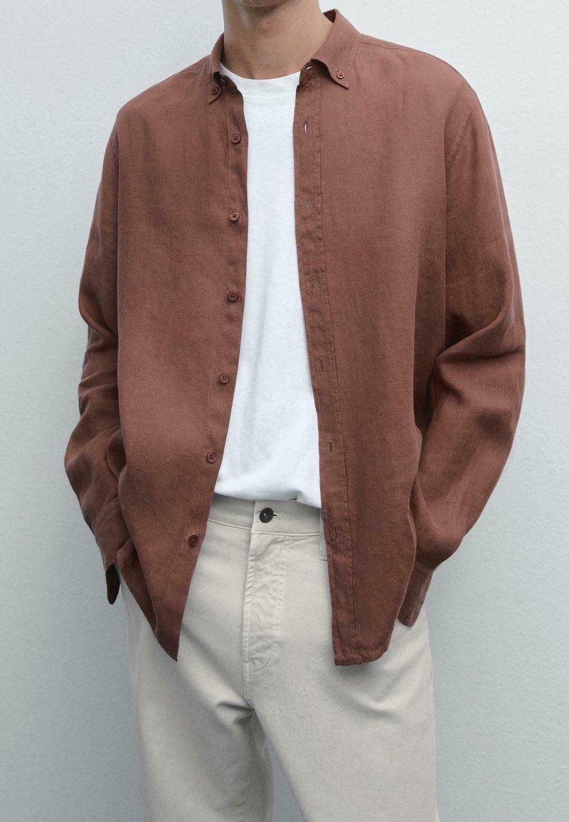 Personne portant une chemise marron à manches longues déboutonnée sur un t-shirt blanc avec un pantalon beige clair, les mains dans les poches.
