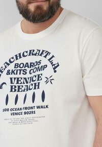 Witte katoenen t-shirt met een grote marineblauwe grafische print met tekst, zon en surfplank iconen, ronde halsontwerp en korte mouwen.