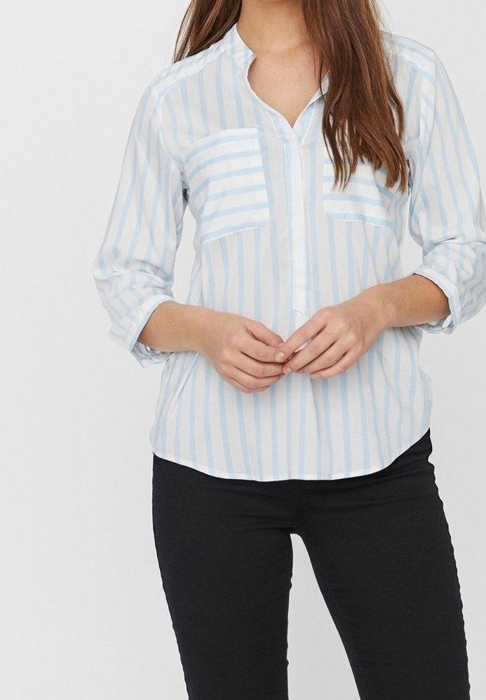 Žena v světle modré a bílé pruhované blůze s dvěma náprsními kapsami a černými kalhotami, stojící na pozadí bez vzoru.