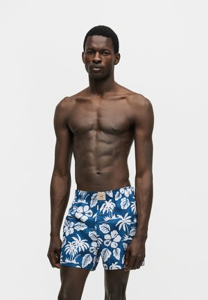 Skjorteløs mand med mørk hud iført blå badeshorts med hvide tropiske blomsterprint, stående mod en ensfarvet lys baggrund.