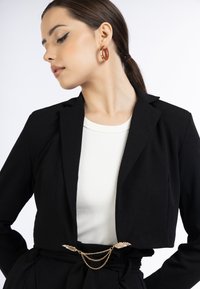 Sort cropped blazer med strukturerede slag, en hvid ribbet top nedenunder, og et guld kæde bælte med bladbunke detaljer. Glat tekstur.