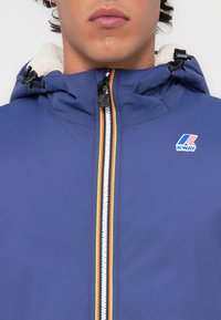 Blå jakke med hette, med en hvit glidelås, rød og gul stripe, og K-Way-logo. Myk fôr synlig i krageområdet.