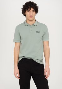 Polo shirt in light green cotton with black trim and EA7 logo. Features a classic collar and short sleeves. Worn with black jeans.