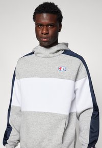 Sudadera gris y blanca con acentos en azul marino, que cuenta con un cuello alto y un logo en el pecho. Fabricada con un tejido suave y un diseño deportivo.
