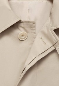 Beige Trenchcoat-Stoff mit einem strukturierten Kragen, ausgestattet mit einem großen Kunststoffknopf und einer dezenten Nähdetails am Revers.