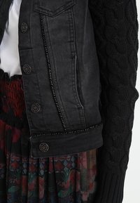 Mustat farkkutakki koristeellisilla napilla mustan lettikäänteen päällä, yhdistettynä värikkääseen, kuvioituun laskoshameeseen ja valkoiseen puseroon.