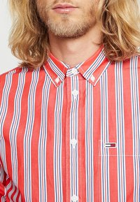 Camicia a righe rosse e bianche con accenti blu, colletto con bottoni e un piccolo logo ricamato sulla tasca del petto.