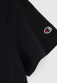 Sort bomulds-T-shirt med korte ærmer, prydet med et lille broderet logo i rød, hvid og grå på ærmet. Glat tekstur.