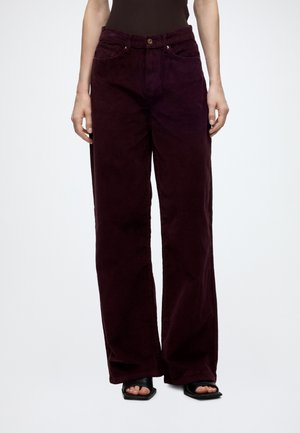 Femme portant un pantalon large en velours côtelé violet foncé avec poches avant et des sandales à talons noires à bout ouvert sur fond blanc.