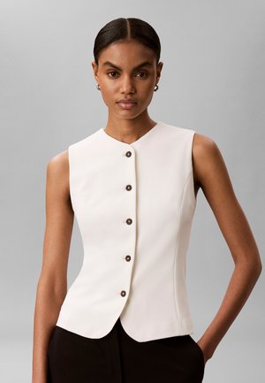 Sötét hajú nő fehér ujjatlan, gombos mellényben és fekete nadrágban, az egyik kezével a zsebében áll egy egyszínű szürke háttér előtt.