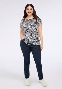 Mujer sonriente con cabello largo y oscuro, que lleva pendientes de aro, una blusa corta de manga con un patrón blanco y azul marino, jeans oscuros y zapatillas deportivas blancas.