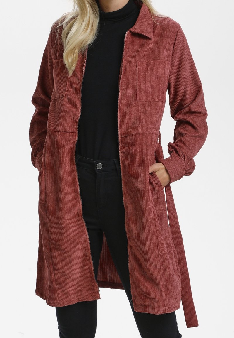 Femme portant un long manteau en velours côtelé couleur rouille avec de grandes poches poitrine, un col roulé noir et un pantalon noir.
