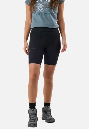 Femme portant un short de randonnée noir à hauteur des genoux, un t-shirt gris à motif, des chaussettes noires et des chaussures de randonnée grises, debout sur un fond blanc.