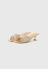 Sandales à talons ouverts dorées avec des lanières métalliques entrecroisées, ornées de strass. Présente un talon courbé et sculpté.