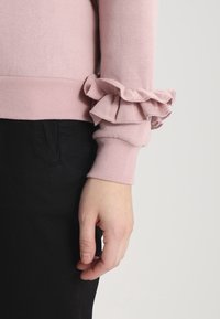 Suéter rosa claro con dobladillo y puños acanalados, adornado con volantes en capas, combinado con pantalones negros. Primer plano del detalle de la manga.