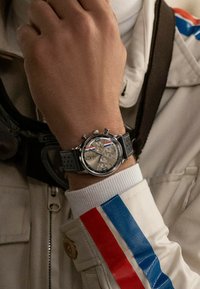 Srebrny zegarek chronograficzny z szarym gumowym paskiem, textured tarczą, trzema sub-tarczami i małą koronką. Posiada biały rękaw z czerwonymi i niebieskimi paskami.