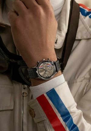 Srebrny zegarek chronograficzny z szarym gumowym paskiem, textured tarczą, trzema sub-tarczami i małą koronką. Posiada biały rękaw z czerwonymi i niebieskimi paskami.