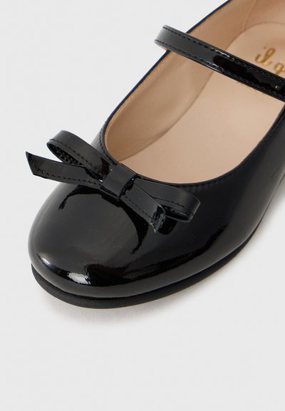 Ballerines en cuir verni noir avec un bout rond, présentant une finition brillante et un accent de nœud décoratif à l'avant, ainsi qu'une seule sangle.