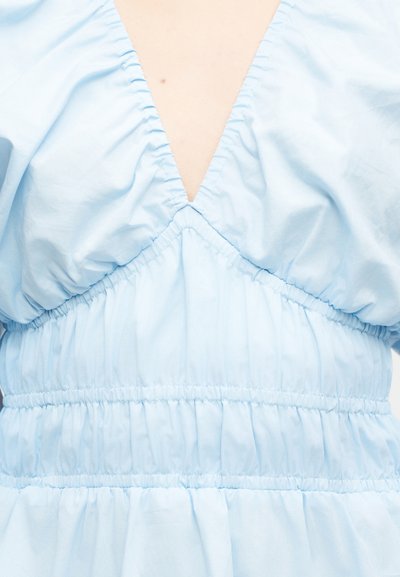 Φωτεινό γαλάζιο φόρεμα με βαθύ V-λαιμόκοψη και συγκεντρωμένες ελαστικές ραφές γύρω από το στήθος και τη μέση σε άτομο με ανοιχτόχρωμο δέρμα.