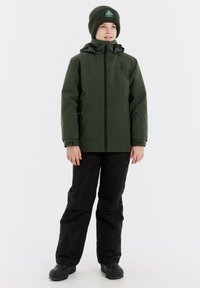 Grüne wasserdichte Jacke mit schwarzen Hosen. Die Jacke hat einen Frontreißverschluss, eine Kapuze und verstellbare Bündchen. Schwarze Winterstiefel vervollständigen das Outfit.