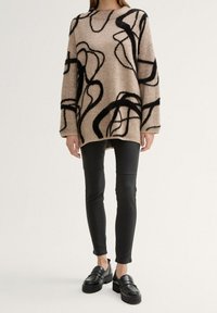 Beige neule, jossa on mustia abstrakteja kuvioita, neulottu pinta, yli suuri malli, yhdistettynä mustiin slim-fittiin housuihin ja kookkaisiin mustiin loafer-kenkiin.