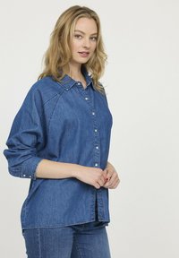 Chemise en denim bleu, avec un devant boutonné, un col, des manches longues avec des poignets ajustables, et une coupe décontractée.